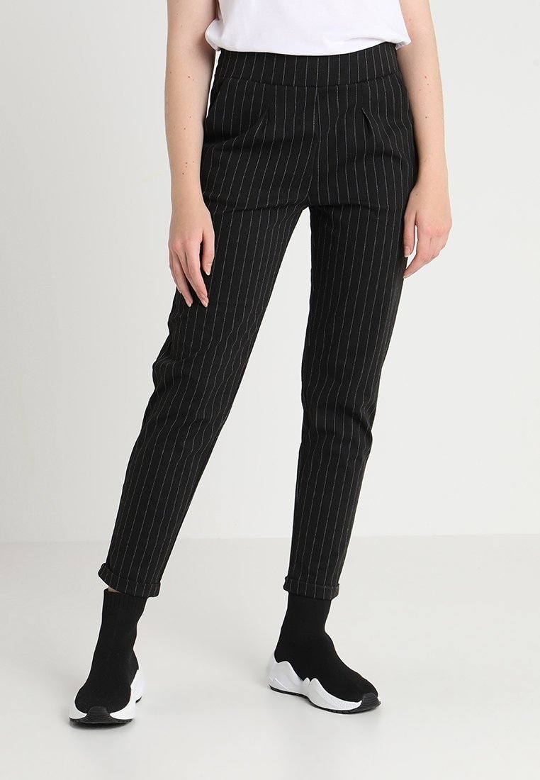 Personne portant un pantalon noir à fines rayures ajusté, des baskets style chaussettes noires avec semelles blanches et un haut blanc sur un fond uni.