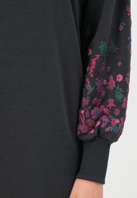 Mørkfarget plagg med lange, puffede ermer og livlige blomsterbroderier i nyanser av rosa, lilla og grønt på mansjettene.
