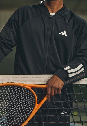 Personne en veste de sport noire avec des bandes blanches tenant une raquette de tennis orange reposant sur un filet de tennis.