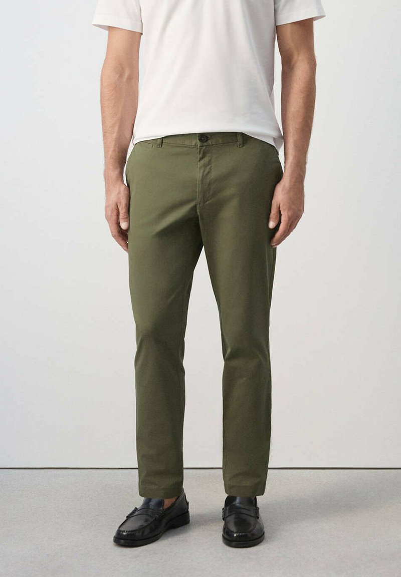 Homme portant un pantalon slim vert olive, une chemise blanche à manches courtes et des mocassins en cuir noirs, debout sur un sol gris.