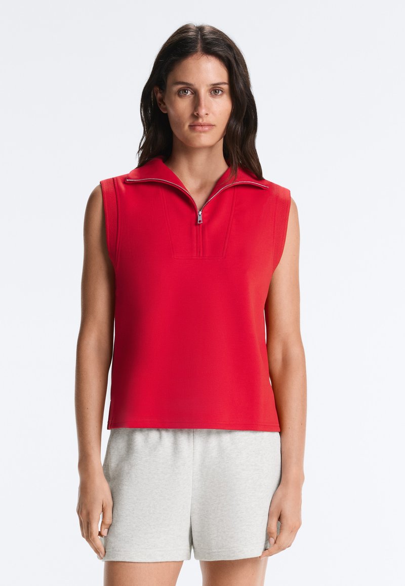 Femme portant un top rouge sans manches zippé et un short gris clair, debout devant un fond clair uni.