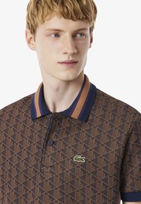 Polo à manches courtes bleu marine avec un motif géométrique marron, doté d'un col bicolore et du logo Lacoste sur la poitrine.