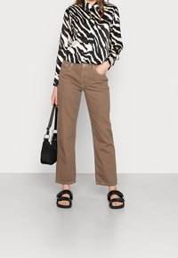 Pantaloni in cotone marroni con vestibilità rilassata, abbinati a una camicia con stampa zebra bianca e nera. Le calzature presentano spesse fasce nere.