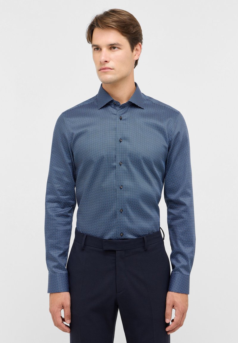 Blau gemustertes Hemd mit Knöpfen, Kragen und langen Ärmeln. Verfügt über ein geometrisches Design und braune Knöpfe. Kombiniert mit dunklen Hosen.