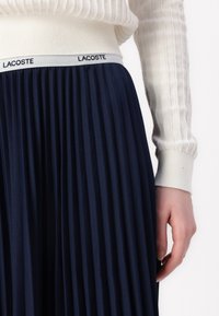 Navy pleated skirt with elastic waist featuring "LACOSTE" branding, paired with a textured cream sweater. Close-up of hand on fabric.