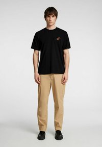 Svart bomullströja med korta ärmar, försedda med en orange logotyp. I kombination med beige byxor och svarta skor står modellen mot en enfärgad bakgrund.