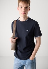 Tamsiai mėlyna marškinė su balta apdaila ir "BOSS" logotipu ant krūtinės, derinami su šviesiai mėlynomis džinsais, laikomi su kreminės spalvos petine rankine.