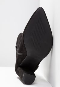 Bottine en suédine noire avec un bout pointu, fermeture éclair sur le côté et une semelle en caoutchouc texturé. Elle présente un talon stable et effilé.