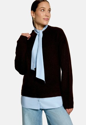Femme portant un pull noir par-dessus une blouse bleu clair avec une cravate assortie, associée à un jean bleu clair, regardant légèrement sur le côté.