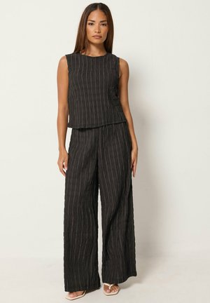Femme portant un haut noir sans manches texturé et un pantalon large, debout devant un fond clair avec une expression neutre.