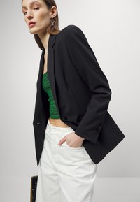Blazer nero con spalle strutturate, chiusura con un bottone, sopra un top a coste verde, abbinato a pantaloni bianchi a vita alta.
