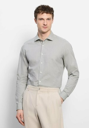 Uomo con capelli corti e ricci che indossa una camicia a righe grigio chiaro a maniche lunghe con bottoni e pantaloni beige, in piedi su sfondo bianco.