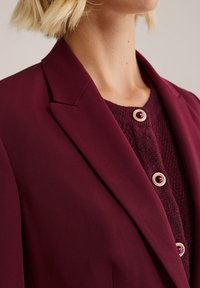 Burgundský blazer s ostrými klopami, doplněný o strukturovaný burgundský svetr se dvěma kulatými knoflíky s zlatými detaily.