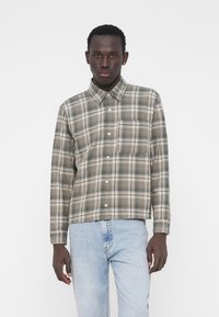 Koszula w kratę w stonowanych odcieniach zieleni i beżu. Posiada standardowy kołnierzyk, długie rękawy oraz kieszeń na piersi. Nosi się ją z jasnoniebieskimi dżinsami.