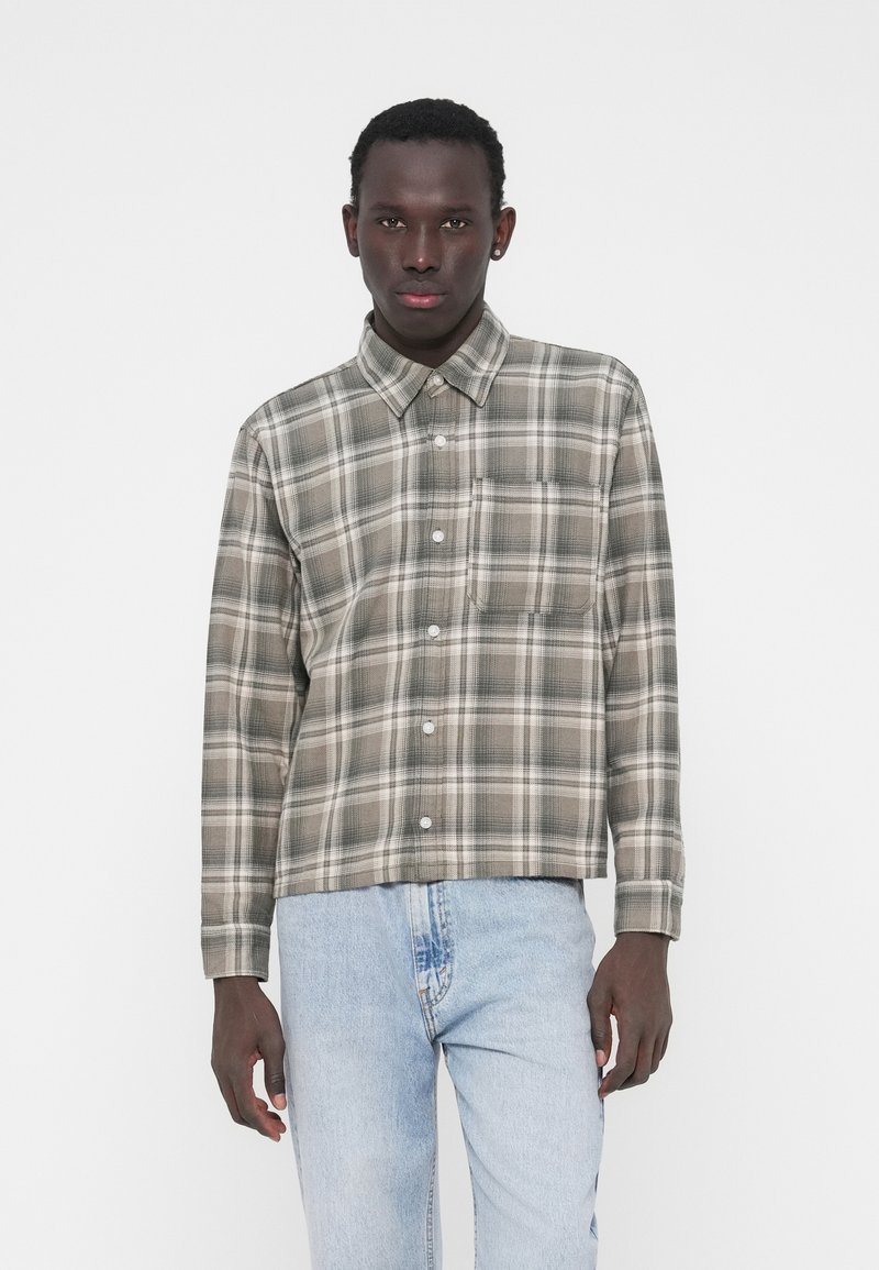 Koszula w kratę w stonowanych odcieniach zieleni i beżu. Posiada standardowy kołnierzyk, długie rękawy oraz kieszeń na piersi. Nosi się ją z jasnoniebieskimi dżinsami.