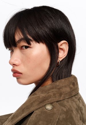 Młoda osoba o prostych czarnych włosach i srebrnej kolczyku kołowym, ubrana w brązową zamszową kurtkę z metalowymi guzikami, patrząca na bok.