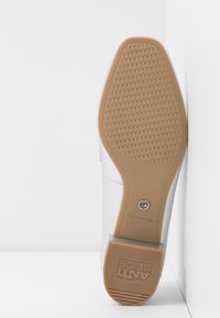 Vue inférieure d'une chaussure blanche avec semelle antidérapante beige, motif de grip texturé et talon bloc, taille 37, posée contre des murs blancs.