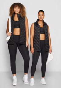 Coletes acolchoados pretos, sem mangas, usados sobre tops curtos pretos, combinados com leggings pretas e ténis brancos. Os coletes apresentam colarinhos altos e fechos de correr.