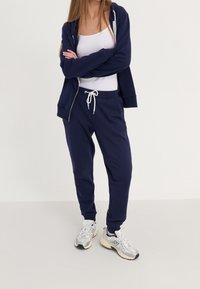 Sudadera con capucha azul marino con cremallera y cordón blanco, combinada con pantalones deportivos a juego en azul marino. Camiseta sin mangas blanca debajo y zapatillas deportivas grises.