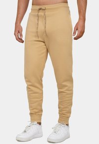 Beige sweatshirtbroek met een elastische tailleband en trekkoord, met gebreide enkels, voorzien van geborduurd merk op de bovenbeen. Gemaakt in combinatie met witte sneakers.