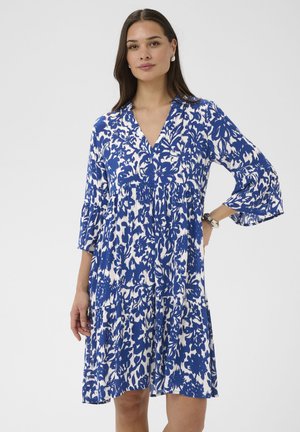 Femme portant une robe mi-longue bleue et blanche à motifs floraux avec des manches cloche 3/4, debout avec une main sur la hanche sur un fond uni.