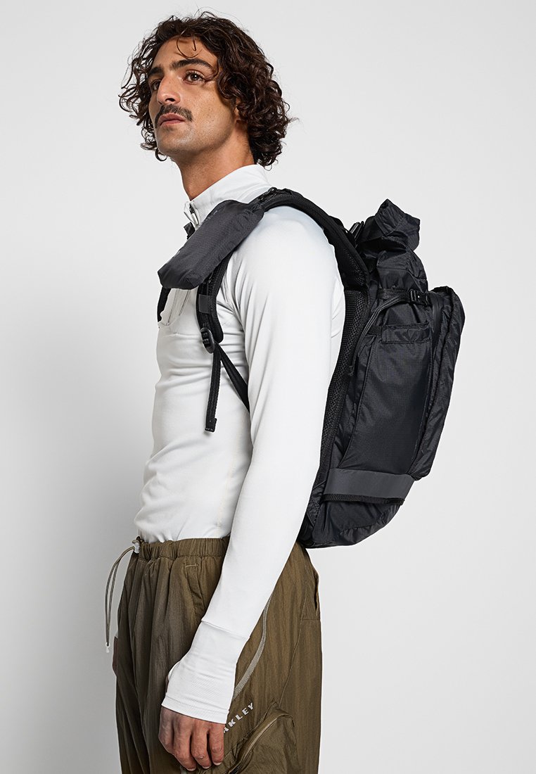Schwarzer Rucksack mit einem strukturierten Design, mehreren Taschen, verstellbaren Schulterriemen und einem Roll-Top-Verschluss. Wird über die Schulter einer Person getragen.