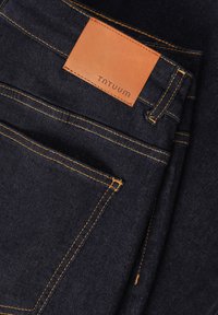 Tmavě modré džíny z denimu s oranžovým prošitím a světlou koženou nášivkou označenou "TNTUUM." Vyznačují se designem s pěti kapsami a různými detaily prošití.