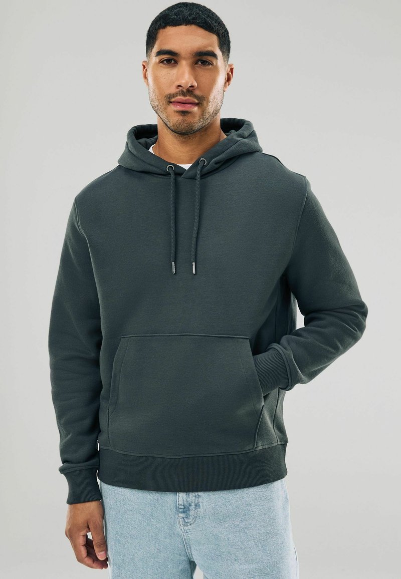 Tmavo zelená kapucňová mikina vyrobená z mäkkej tkaniny, s prednou vreckom, rebrovanými manžetami a voľným strihom.