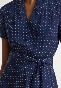 Nærbilde av marineblå kjole med hvite prikker, korte ermer, knappet front og knute i midjen laget av stoffet på en person.