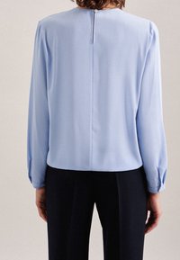 Blusa a maniche lunghe blu chiaro con scollatura rotonda, zip sul retro e dettagli a bottone sui polsini. Tessuto morbido con una caduta delicata.