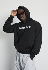 Black hoodie with a front pocket, featuring white text "forgotten faces" in a stylized font. Model wears sunglasses and light gray pants.