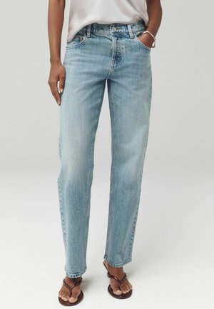 Person trägt hellblaue Straight-Leg-Jeans, weißes Top, braune Sandalen, mit einer Hand in der Tasche und einem silbernen Armband am Handgelenk.
