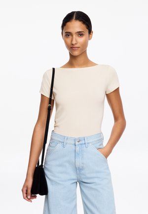 Frau trägt ein beiges Kurzarmoberteil und hellblaue Jeans mit hoher Taille, hält eine schwarze Schultertasche und hat eine Hand in der Tasche.