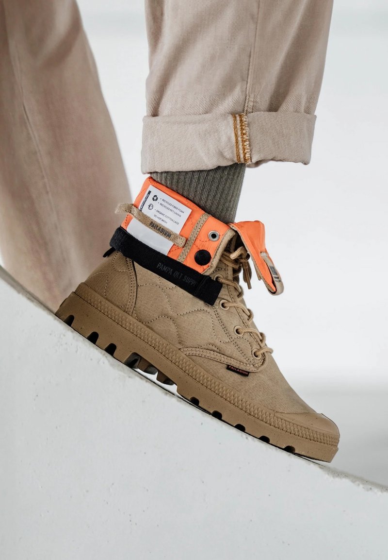 Bottine montante en toile couleur kaki clair avec accents orange et noir, dessus texturé, semelle en caoutchouc et détail du logo visible sur la languette et le côté.