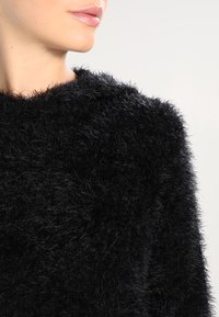 Schwarzer, flauschiger Pullover mit weicher Textur, rundem Halsausschnitt und langen Ärmeln. Das Material wirkt plüschig und leicht glänzend im Licht.