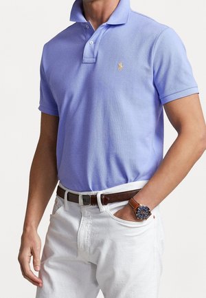 Mężczyzna ubrany w jasno fioletową koszulkę polo z logo, białe spodnie, brązowy skórzany pasek oraz zegarek na rękę z niebieską tarczą i brązowym paskiem.