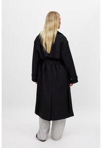 Cappotto lungo nero con vita abbinata, ampi revers e polsini abbottonati. Materiale liscio e lucido, lunghezza fino al ginocchio con spacco posteriore.