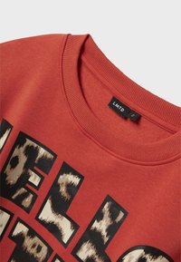 Sudadera roja con cuello redondo acanalado, gran texto negro y un gráfico de estampado de leopardo en tonos de beige y marrón. Etiqueta de la marca visible.