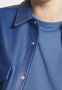 Blauwe leren jas met een shirtkraag, voorzien van zilveren drukkers en een gladde textuur, gelaagd over een blauwe t-shirt met ronde hals.