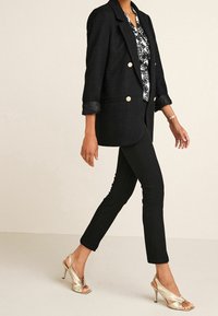 Femme marchant avec un pantalon noir court, un blazer noir à boutons dorés, un chemisier à motifs et des sandales à talons métalliques.