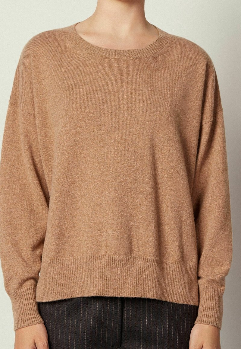 Personne portant un pull ample en maille de couleur camel avec des poignets et un ourlet côtelés, assorti à un pantalon sombre à fines rayures.
