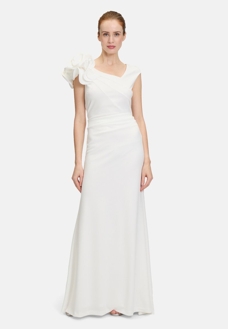 Robe blanche sans manches avec un corsage ajusté, décolleté en V, et un grand accent floral sur l'épaule gauche. Tissu lisse et fluide allant jusqu'au sol.