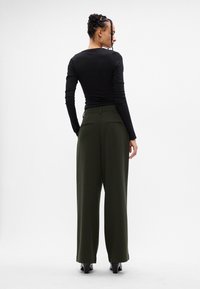 Γυναίκα με πλεξούδα, φορώντας ένα μαύρο μπλουζάκι μακρύ με μανίκια, σκούρους πράσινους φαρδιούς παντελόνες και μαύρα τακούνια, γυρτή προς τα πίσω σε λευκό φόντο.