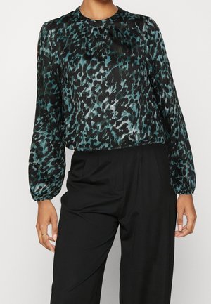 Kobieta w długim rękawie bluzce w turkusowo-czarny zwierzęcy wzór, włożonej do wysokich czarnych spodni, stojąca z rozluźnionymi ramionami.