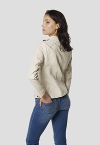 Beige Lederjacke mit figurbetontem Schnitt und Nahtdetails, kombiniert mit blauen Jeans mit Ziernähten und Gesäßtaschen.