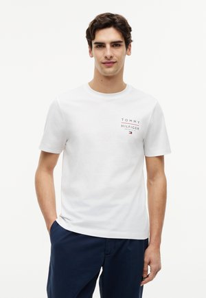 Νέος άνδρας που φοράει λευκό μπλουζάκι Tommy Hilfiger και σκούρο μπλε παντελόνι, στέκεται με το ένα χέρι στην τσέπη του μπροστά σε απλό φόντο.