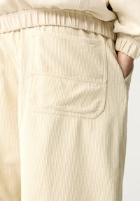 Beige corduroy broek met getextureerde verticale strepen, voorzien van een elastische tailleband en een paspelzak aan de zijkant.