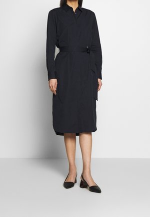 Vrouw draagt een zwarte midi blousejurk met lange mouwen en ceintuur en zwarte schoenen met lage blokhak, staand tegen een effen achtergrond.