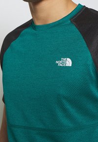 Sportowa koszulka w kolorze niebiesko-zielonym z czarnymi akcentami, teksturowanym materiałem, okrągłym dekoltem i logo na lewej piersi. Zaprojektowana dla wygody i swobody ruchów.