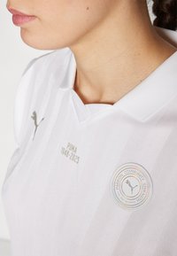 Bijela sportska polo košulja s vertikalnim trakama, PUMA brendiranjem u sivoj boji i kružnim grbom koji sadrži natpis "Strast Zajednica Snaga" i logo.